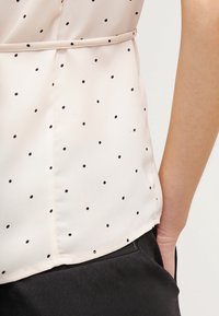 Blusa beige con pois neri infilata in pantaloni neri, che mostra la cucitura laterale e una mano appoggiata nella tasca.