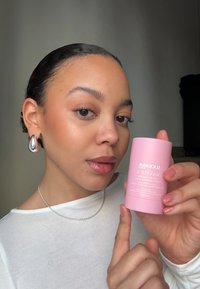 Contenitore cilindrico rosa etichettato "Slick Hair Wax Stick" in testo bianco. Finitura opaca liscia, tenuto da una persona con una collana di catena argentata.