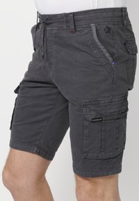 Mörkgråa herrcargo-shorts med flera fickor, snörning i midjan och strukturerad tygbärs av en stående person.