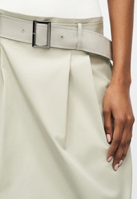 Χέρι που ακουμπάει σε ανοιχτό μπεζ φούστα με πτυχώσεις και φαρδιά ζώνη με ορθογώνιο μαύρο πόμολο.