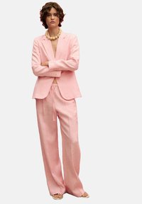 Light pink linen suit consisting of a tailored blazer and wide-leg trousers, paired with a natural bead necklace and tan sandals.