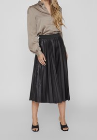 Blusa in satin beige, maniche lunghe, gonna midi nera plissettata, vita elastica e sandali neri aperti. Consistenza liscia in tutto.