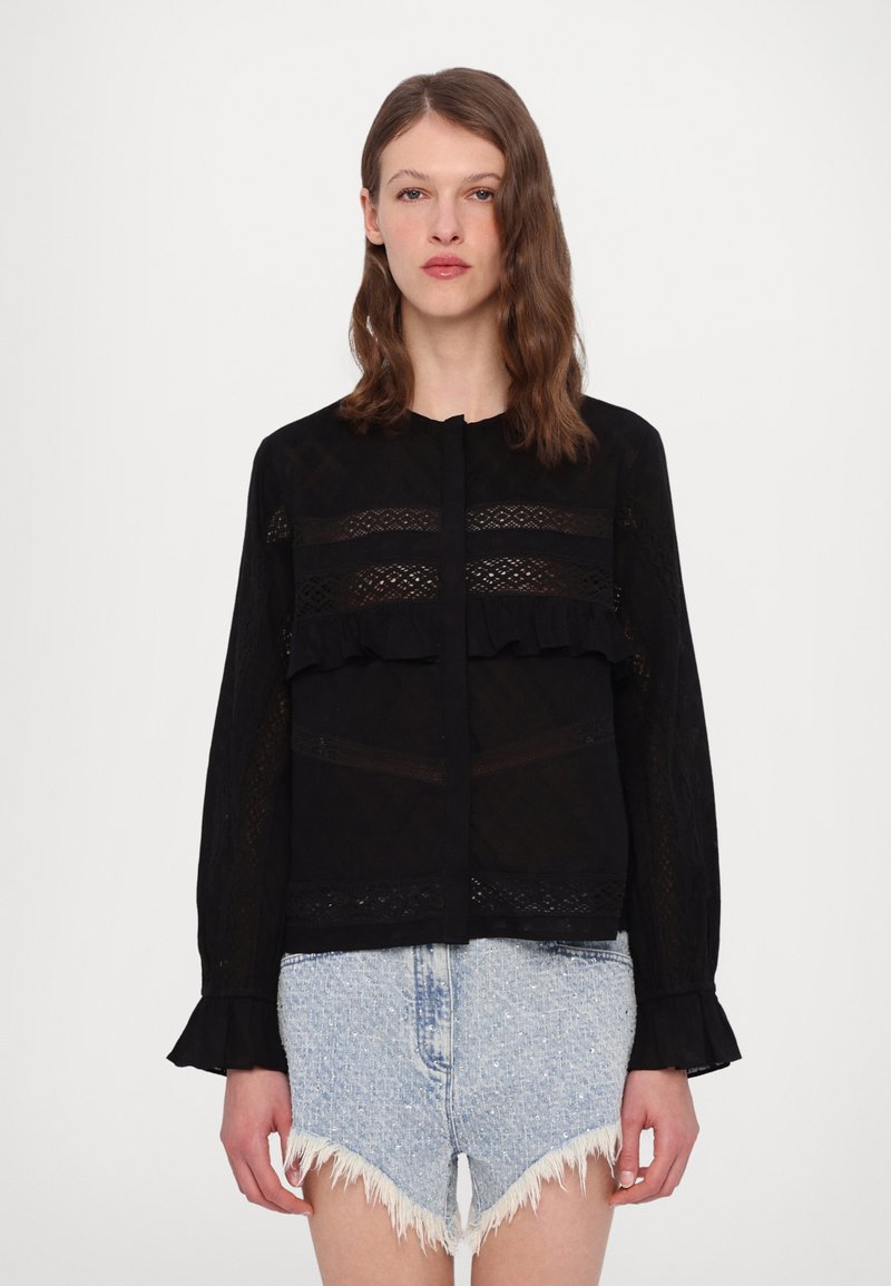 Schwarze Bluse mit Spitze und Rüschendetails, Rundhalsausschnitt und langen Ärmeln. Über hellen Jeansshorts getragen.