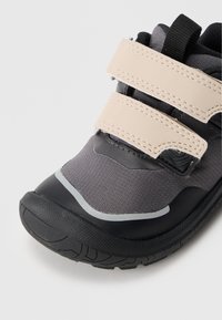 Sapatilha desportiva com parte superior em tecido cinza e detalhes em sintético preto, duas faixas de velcro bege e sola de borracha texturizada para maior aderência.