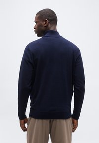Homme vu de dos portant un pull à col roulé bleu marine et un pantalon marron clair sur un fond blanc uni.