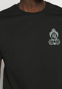 Černé bavlněné tričko s kulatým výstřihem a krátkými rukávy, s designem světlých květin a symbolů na hrudi.