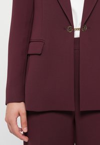 Bordeaux op maat gemaakte blazer met een gestructureerde pasvorm, voorzien van een borstzak, dubbele knopen en een kettingaccent bij de sluiting. Gladde stoftextuur.