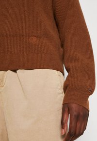 Brauner gerippter Pullover mit strukturierter Oberfläche und kleinem Logo-Emblem. Kombiniert mit hellbeigen Hosen mit seitlichen Taschen.