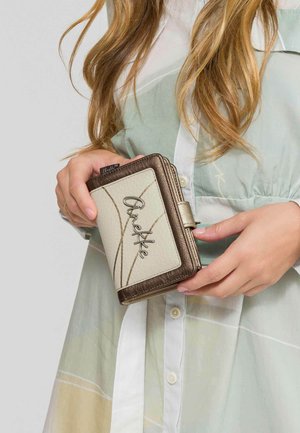 Mujer sosteniendo una cartera texturizada beige y bronce con la palabra "Anekke" en el frente, vistiendo un vestido abotonado de colores pastel.