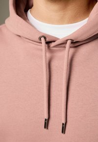 Sudadera de color rosa claro con cordón y puntas de metal plateado, tejido texturizado y una camiseta blanca superpuesta.