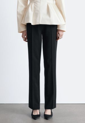 Person iført sorte, højtaljede bukser med lige ben, spidse sorte sko og en cremefarvet peplum-bluse, stående på en grå gulv.