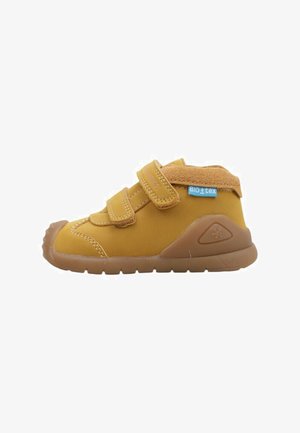Chaussure jaune pour enfants avec deux sangles Velcro, semelles en caoutchouc et un capuchon de orteil texturé en beige. Étiquette "BIO_tex" visible sur le côté.