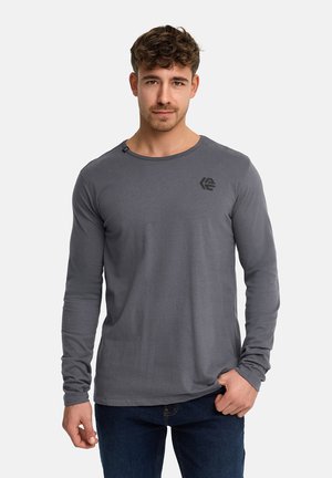 Junger Mann mit kurzen lockigen Haaren, der ein eng anliegendes dunkelgraues Langarmshirt und dunkelblaue Jeans trägt, vor einem schlichten Hintergrund stehend.