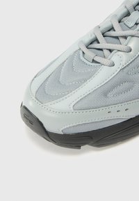 Lichtgrijze sneaker met mesh- en leermaterialen, textuur overlays, vetersysteem en een zwarte rubberen zool met subtiele merkje-details.