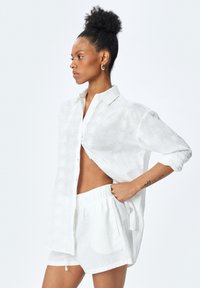 Hvid, letvægts hørskjorte med palmebladmønster, åbnet i siden, parret med matchende shorts. Blød tekstur, afslappet pasform.