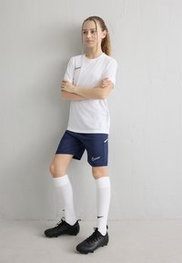 Pusaudze meitene baltā futbola kreklā, tumši zilās šortos, baltās, ceļgalu augstumā garās zeķēs un melnās futbola kedās stāv ar sakrustotām rokām pie baltas sienas.