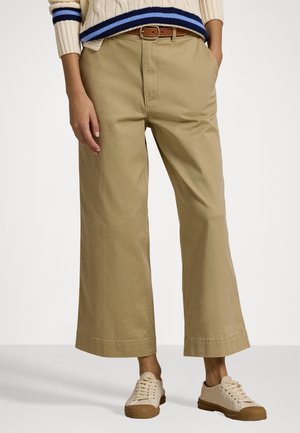 Personne portant un pantalon beige à jambes larges, une ceinture marron, des baskets crème, et un pull crème avec des rayures bleues et noires aux ourlets et aux poignets.