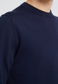 Námořnická modrá pletená svetr s kulatým výstřihem, vyrobený z měkké látky. Detailní záběr ukazuje hladkou texturu a jemně žebrovaný výstřih.