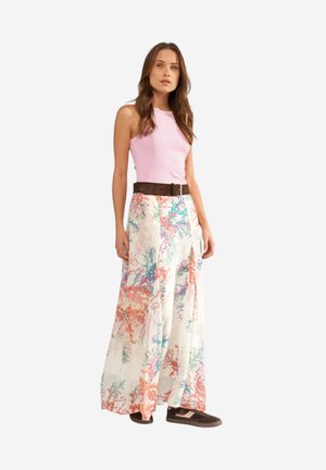 Femme portant un haut rose sans manches, une jupe longue à motif floral avec une ceinture marron et des baskets marron, debout devant un fond blanc.