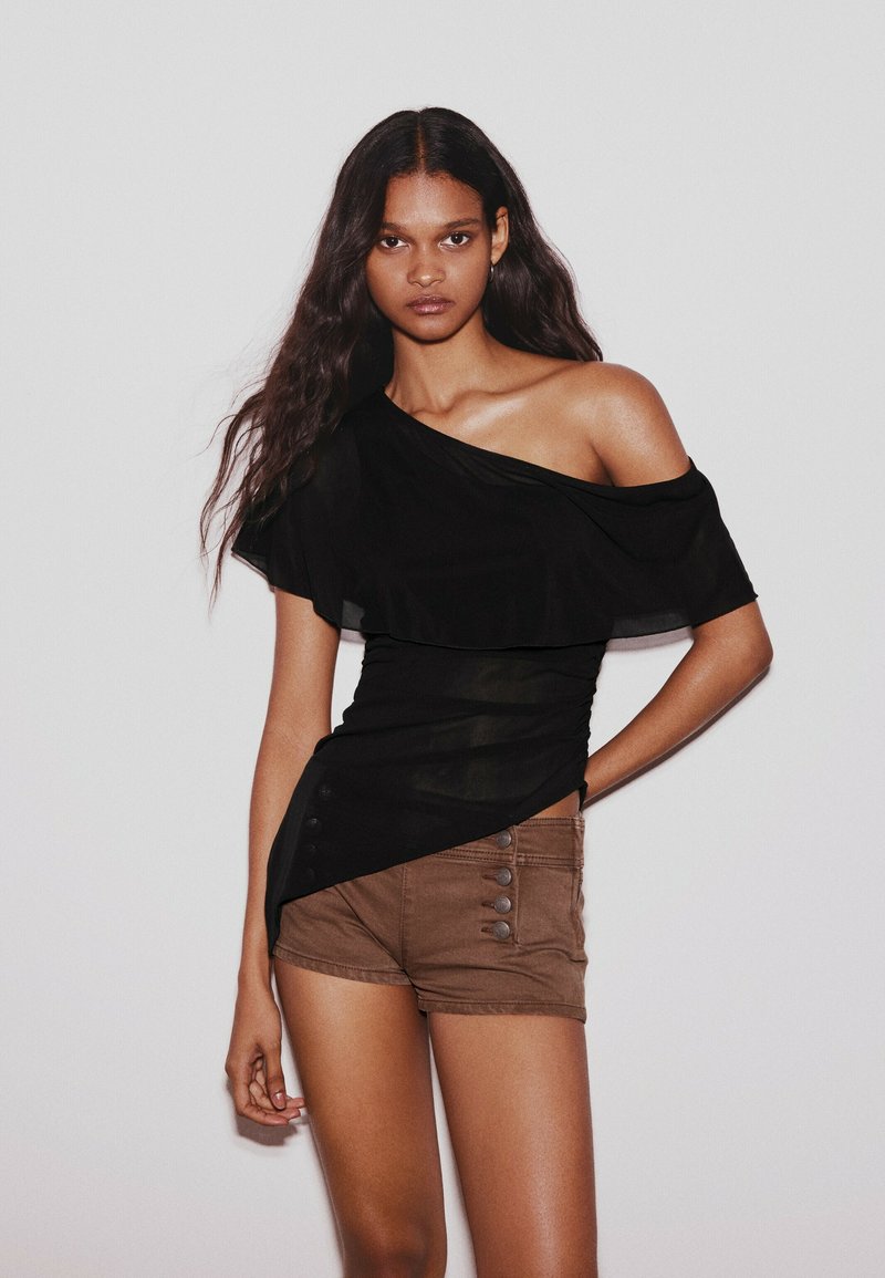 Femme aux longs cheveux ondulés portant un top noir asymétrique et un short marron taille haute sur un fond clair uni.
