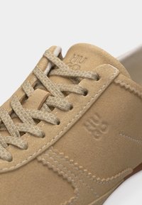 Μπεζ καμηλό δερμάτινο αθλητικό παπούτσι με ομοιόχρωμους κορδόνια, ορατές ραφές και ανάγλυφο λογότυπο "HUCO" στο πλάι και στη γλώσσα.