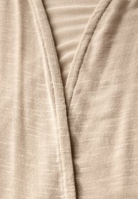 Beige stickad tyg med en subtil textur och ljusa diagonala ränder, som har en djup V-ringning och synliga sömmar.