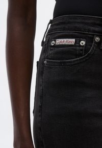 Jeans de mezclilla negra con herrajes plateados, presentan un parche con el logo de Calvin Klein y costuras visibles en los bolsillos.
