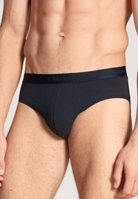 Mannelijk bovenlijf in zwarte Calida slips met een merkband, toont de buik, heupen en bovenbenen tegen een neutrale achtergrond.