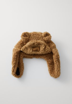 Bonnet en polaire marron conçu avec des accents en forme d'oreilles, des sangles sous le menton et un petit patch logo. Texture douce avec une apparence chaleureuse et confortable.
