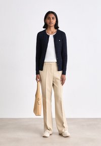 Navy cable-knit cardigan over a white top, beige tailored trousers, and cream slip-on shoes. Holding a tan handbag with a tag.