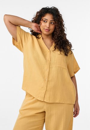 Lokkis juustega naine, kes kannab lahtist kollast nööbitavat särk ja sobivaid pükse, poseerides ühe käega kaela taga.