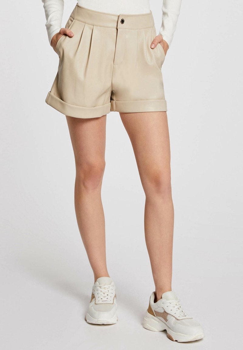 Beige skládací kraťasy s ohrnutým lemem, obsahující boční kapsy a přední zapínání na knoflík, vyrobené z hladkého a lehkého materiálu.