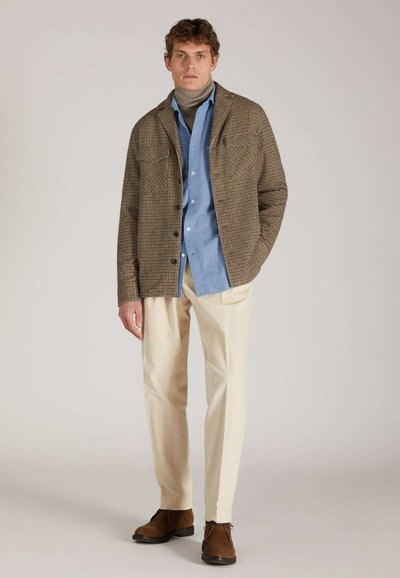 Giacca a quadri marrone e crema sopra una camicia azzurro chiaro, abbinata a pantaloni beige e scarpe in suede marroni. Due tasche sul petto visibili.