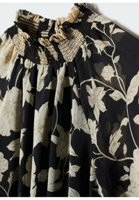 Blouse noire à imprimé floral beige, avec un col haut froncé smocké et un tissu froncé sous le col.