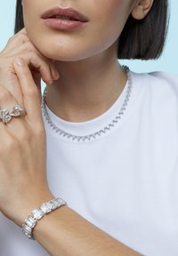 Sølv smykker med en trekantet halskæde, et rektangulært armbånd og en ring med ovale accenter, alt sammen sat mod en hvid skjorte.