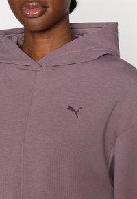 Fialová mikina vyrobená z mäkkého materiálu. Má predné vrecko a malé tmavé logo na hrudi. Textúra sa zdá byť hladká a ľahká.