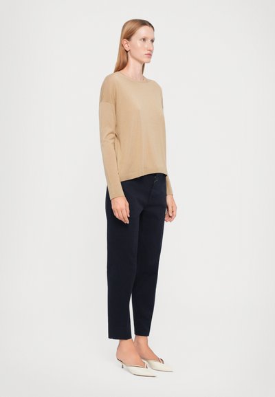 Tan long-sleeve top with a relaxed fit, paired with navy tapered trousers. Model wears pointed white slip-on shoes. Minimalist design, smooth texture.