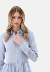 Hellblaues, langärmliges Hemdkleid mit geraffter Taille, aus strukturiertem Stoff. Verfügt über einen spitzen Kragen und Knöpfe vorne.