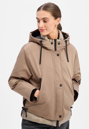 Woman with brown hair tied back wears a beige hooded puffer jacket with black drawstrings and hands in pockets, looking slightly down and right.