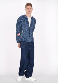 Marineblå zip-up jakke med et spraglet mønster, der har to forlommer og en hætte, parret med løstsiddende blå jeans og hvide sneakers.