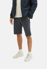 Marineblauer gesteppter Mantel, leichtes Hemd, gemusterte marineblaue Shorts und weiße Sneakers mit grauen Akzenten. Die Shorts haben einen geraden Schnitt und Taschen.