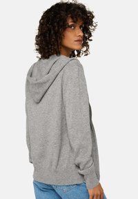 Haut zippé gris en tissu doux ; doté d'une capuche, de poignets côtelés et d'une taille côtelée. Porté avec un jean en denim bleu clair.