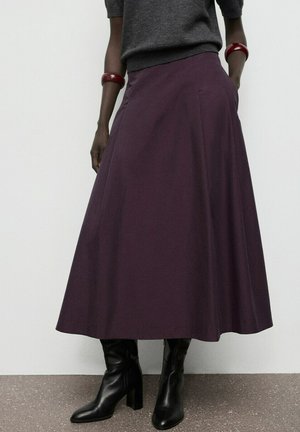 Frau trägt einen dunkelvioletten Midirock, einen anthrazitfarbenen Pullover, schwarze Stiefel mit Absatz und rote Armreifen an beiden Handgelenken, steht mit den Händen in den Taschen.