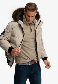 Beżowa kurtka puchowa z kapturem wykończonym futrem, z zamkiem błyskawicznym i czarnymi mankietami, noszona na beżowym fleece i koszuli w kratę, w połączeniu z niebieskimi dżinsami.