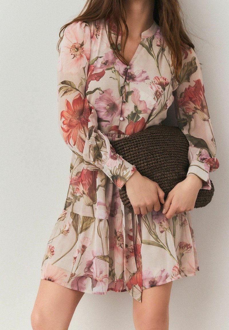 Femme en robe à manches longues à motif fleuri tenant une grande pochette tressée marron foncé sur un fond uni.
