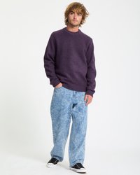 Ung mand med ustyrligt hår iført en lilla sweater, lyseblå acid-wash jeans og sorte lavt-top sneakers, stående med den ene hånd i lommen.