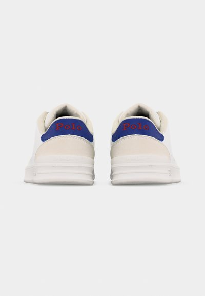 Ζευγάρι λευκά αθλητικά παπούτσια με μπλε ταπί πτέρνας και κόκκινο κείμενο "Polo", φαίνονται από πίσω σε απλό φωτεινό γκρι φόντο.