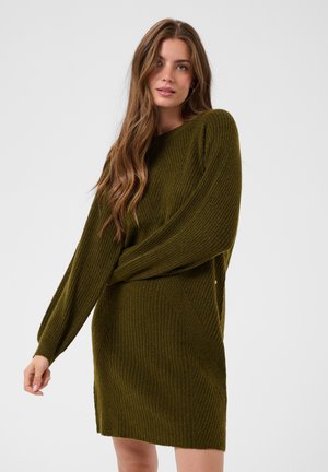 Femme aux longs cheveux bruns portant une robe-pull en maille côtelée vert olive, debout devant un fond clair uni.