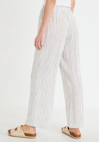 Persona con pantalones blancos de pierna ancha con rayas verticales negras y sandalias beige de doble tira, de pie contra un fondo blanco liso.