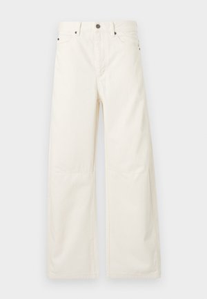 Vrouw met lang donker haar, gekleed in een mouwloos spijkertopje met een tailleriem, wijde witte broek en zwarte puntige hakken, staand tegen een witte achtergrond.